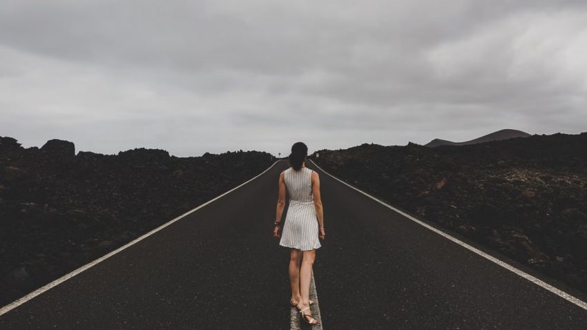 A woman in a white dress walks on an open road surrounded by mountainous landscape.
